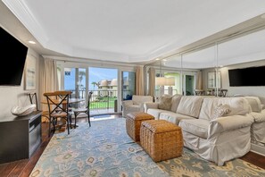 A modern living room with a sofa, ottomans, a TV, and a dining area with chairs and a table. - Watch the waves roll in from every corner. W/D in condo. Upgraded to hardwood. (St. Augustine)