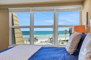 A room with a large window overlooking a beach, a bed with blue bedding, and a lamp on the nightstand.