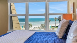 Ein Schlafzimmer mit großem Fenster mit Blick auf einen Strand, einem Bett mit blau-weißer Bettwäsche und einer einzelnen orangefarbenen Nachttischlampe.
