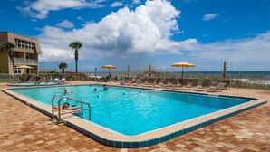 Ein Swimmingpool mit Liegestühlen und Sonnenschirmen, umzäunt, mit einem Gebäude im Hintergrund.
