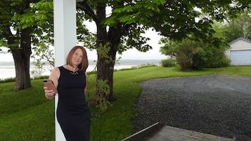 A woman standing on a porch with a view of a lake and a gravel path.