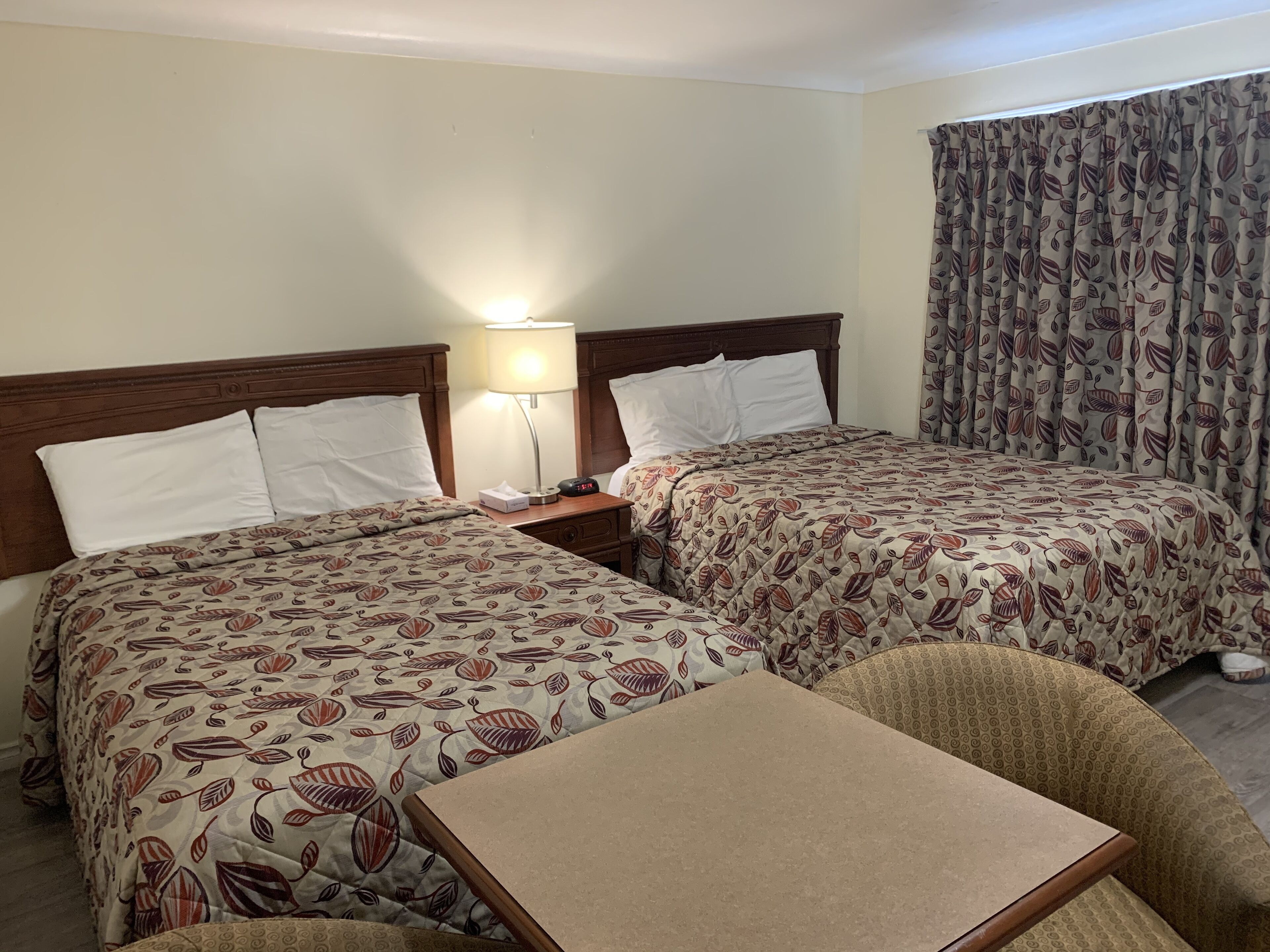 Habitación de hotel con dos camas, una mesita de noche con lámpara, una pequeña mesa y una cortina estampada.