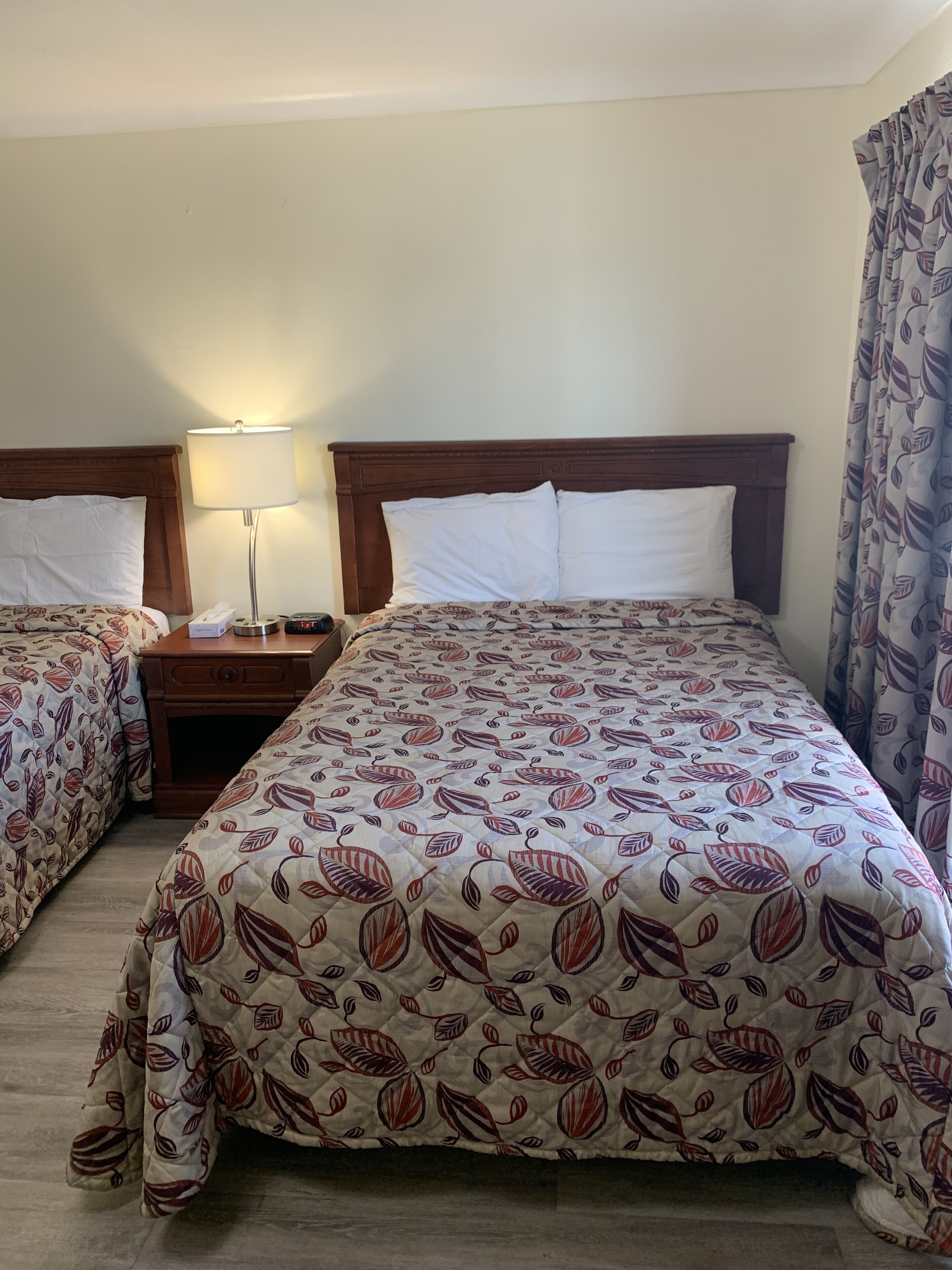 A hotel room with two beds, a nightstand, a lamp, and a curtain.