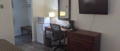 A hotel room with a bed, a desk with a chair, a microwave, a refrigerator, a lamp, a mirror, and a TV.