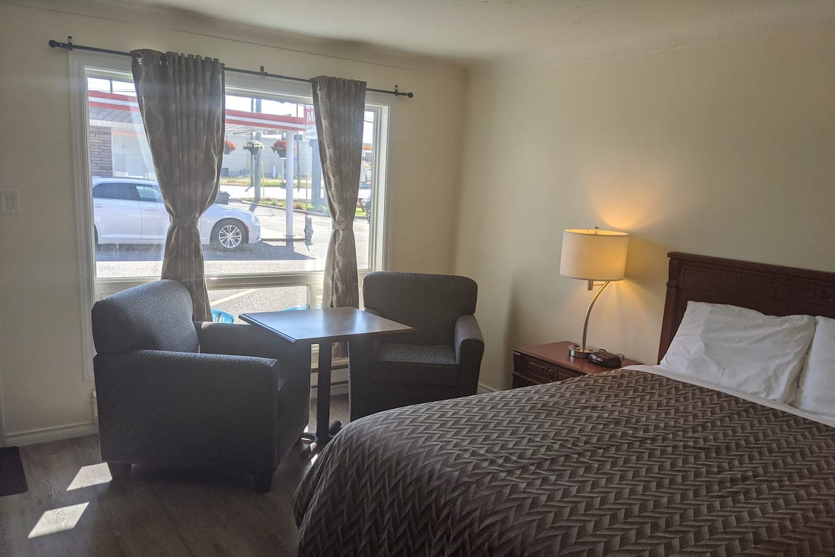A hotel room with a bed, two armchairs, a small table, a nightstand with a lamp, and a window with curtains.