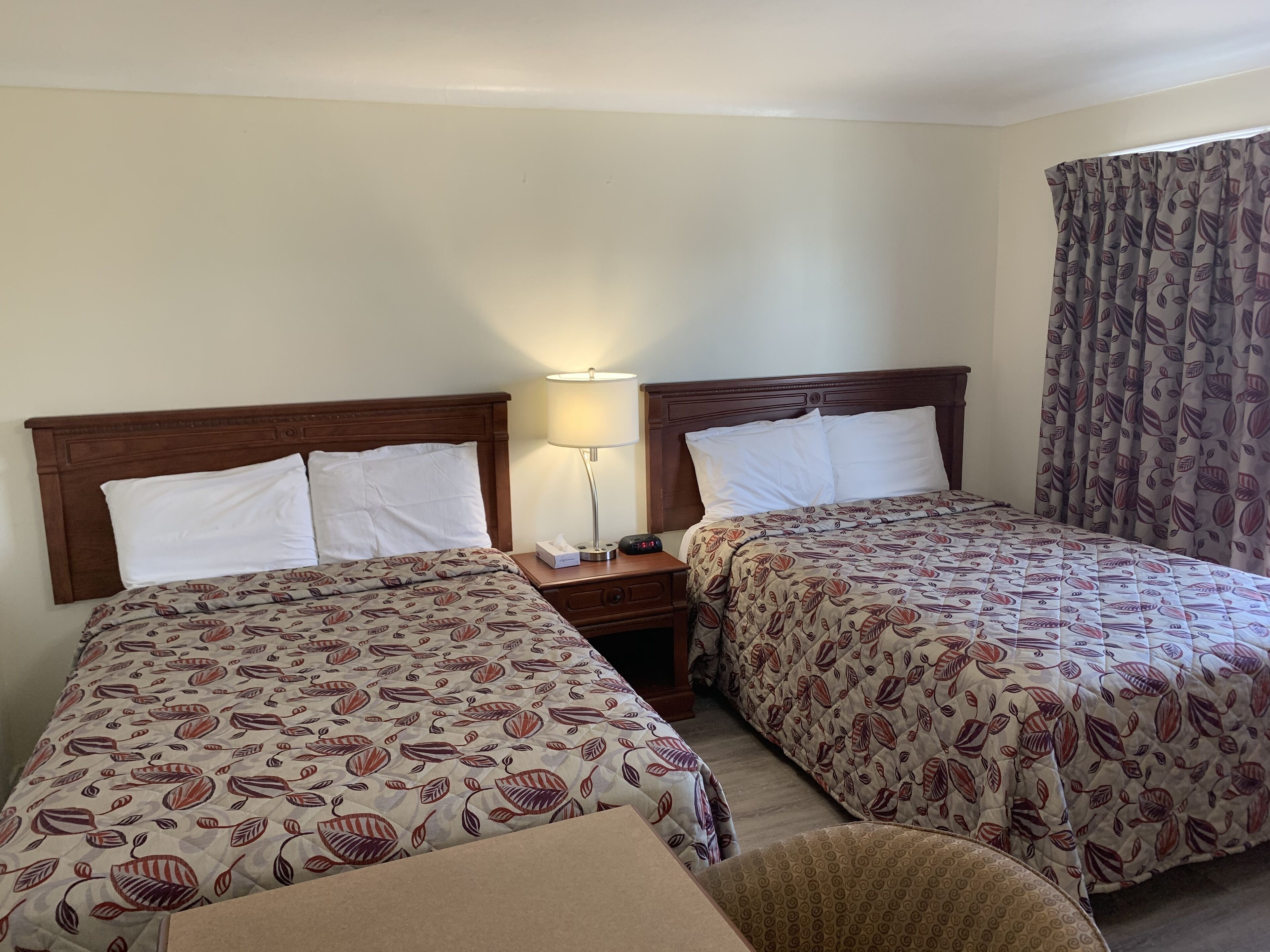 Habitación de hotel con dos camas, una mesita de noche con lámpara y una ventana con cortinas estampadas.