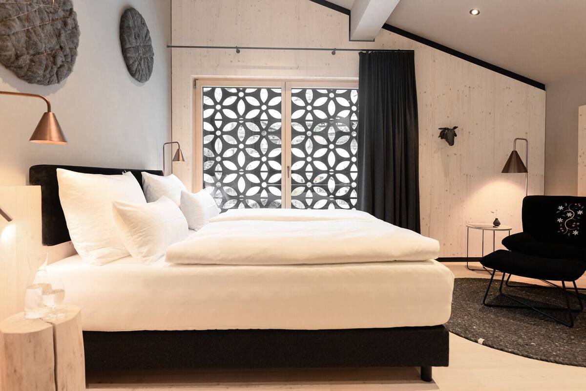 A modern bedroom with a large bed, a black chair, and a decorative window with a geometric pattern.