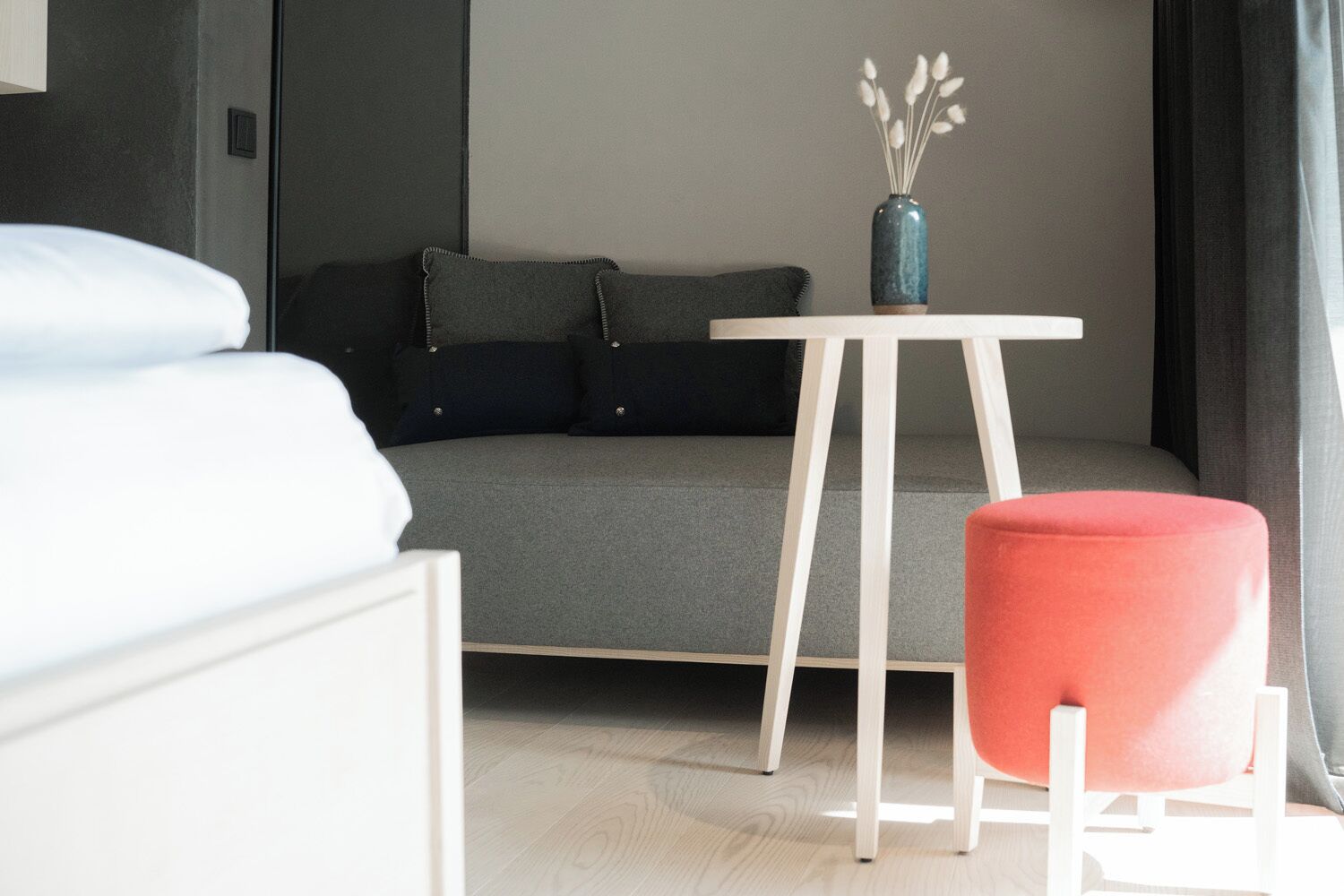 A modern hotel room with a bed, a grey sofa, a small table with a vase, and a red ottoman.