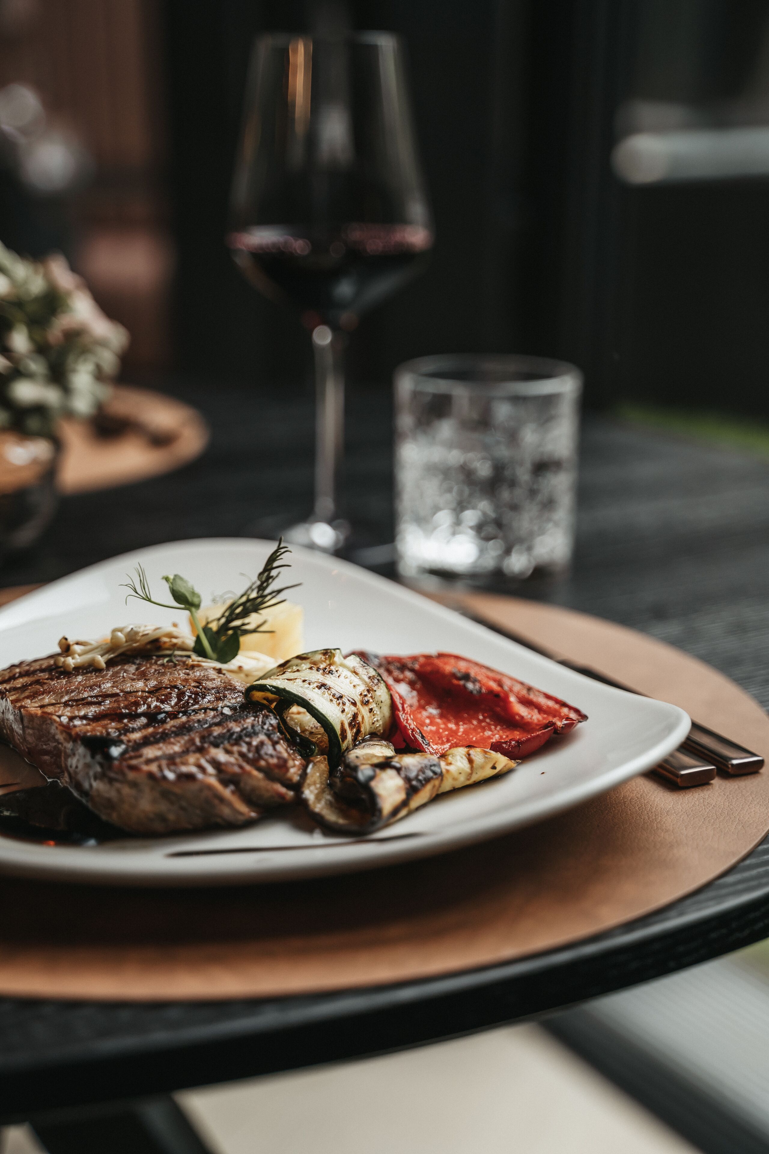 A plate of food with a steak, vegetables, and a garnish on a table with a glass of red wine and a glass of water.