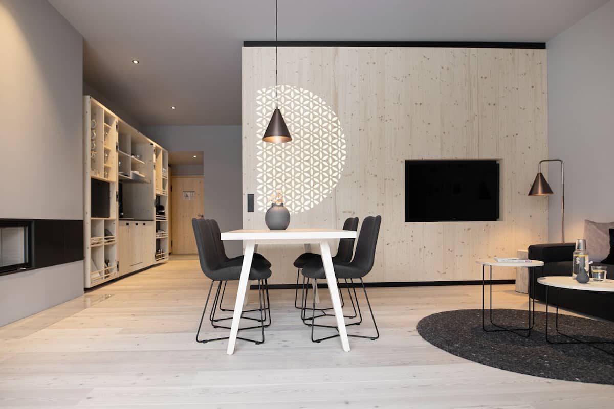 A modern interior with a dining area featuring a white table and black chairs, a black TV mounted on the wall, and a wooden panel with a geometric pattern.
