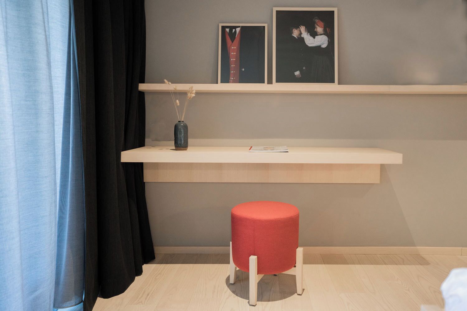 A room with a light wooden floor, a red stool, a shelf with framed pictures, and a vase with dried flowers.
