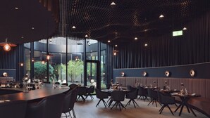 A modern restaurant with a central bar, multiple dining tables, and a view of greenery outside. - Amrai Suites (Schruns)