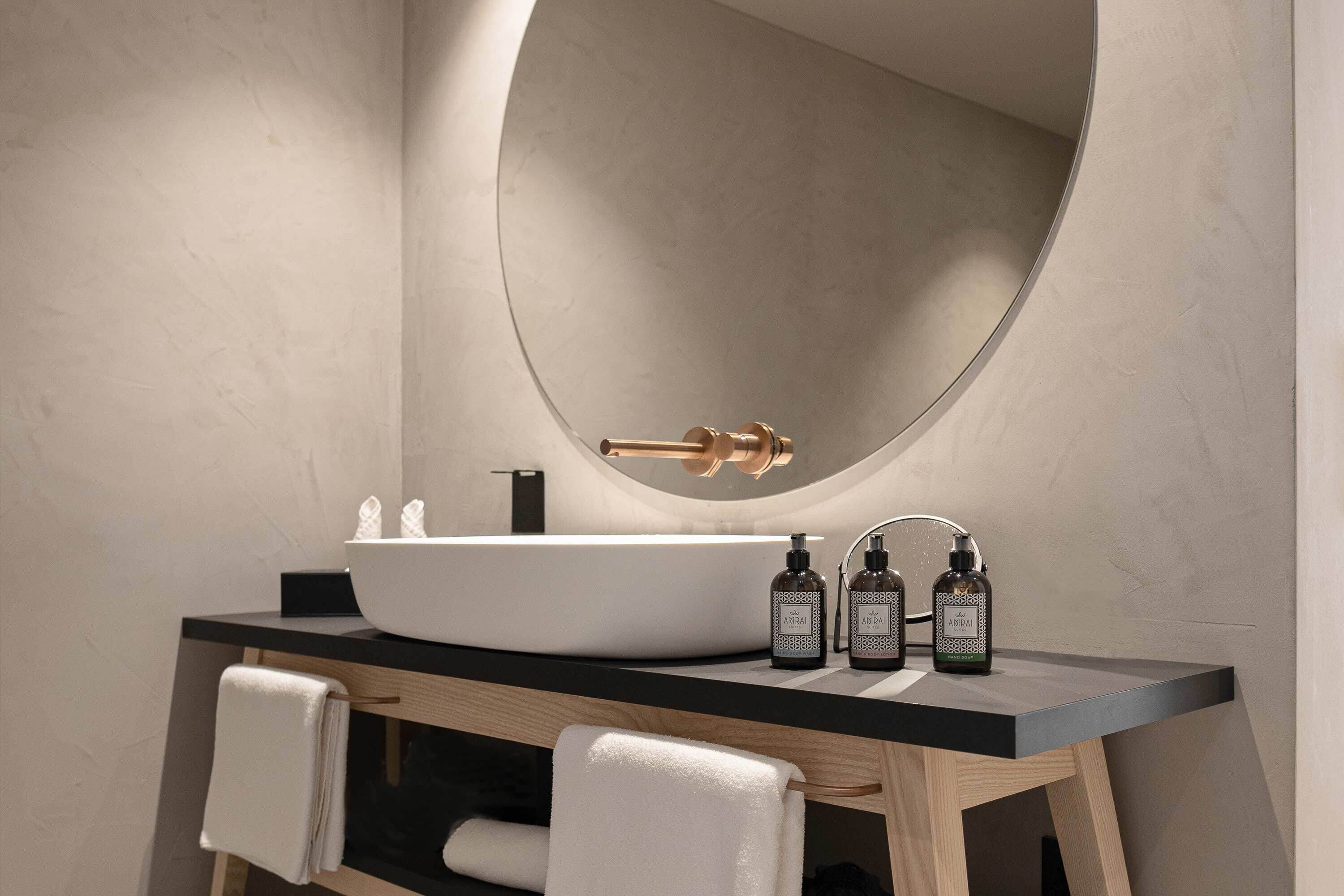 A modern bathroom with a large oval mirror, a white sink, and three bottles of hand sanitizer.