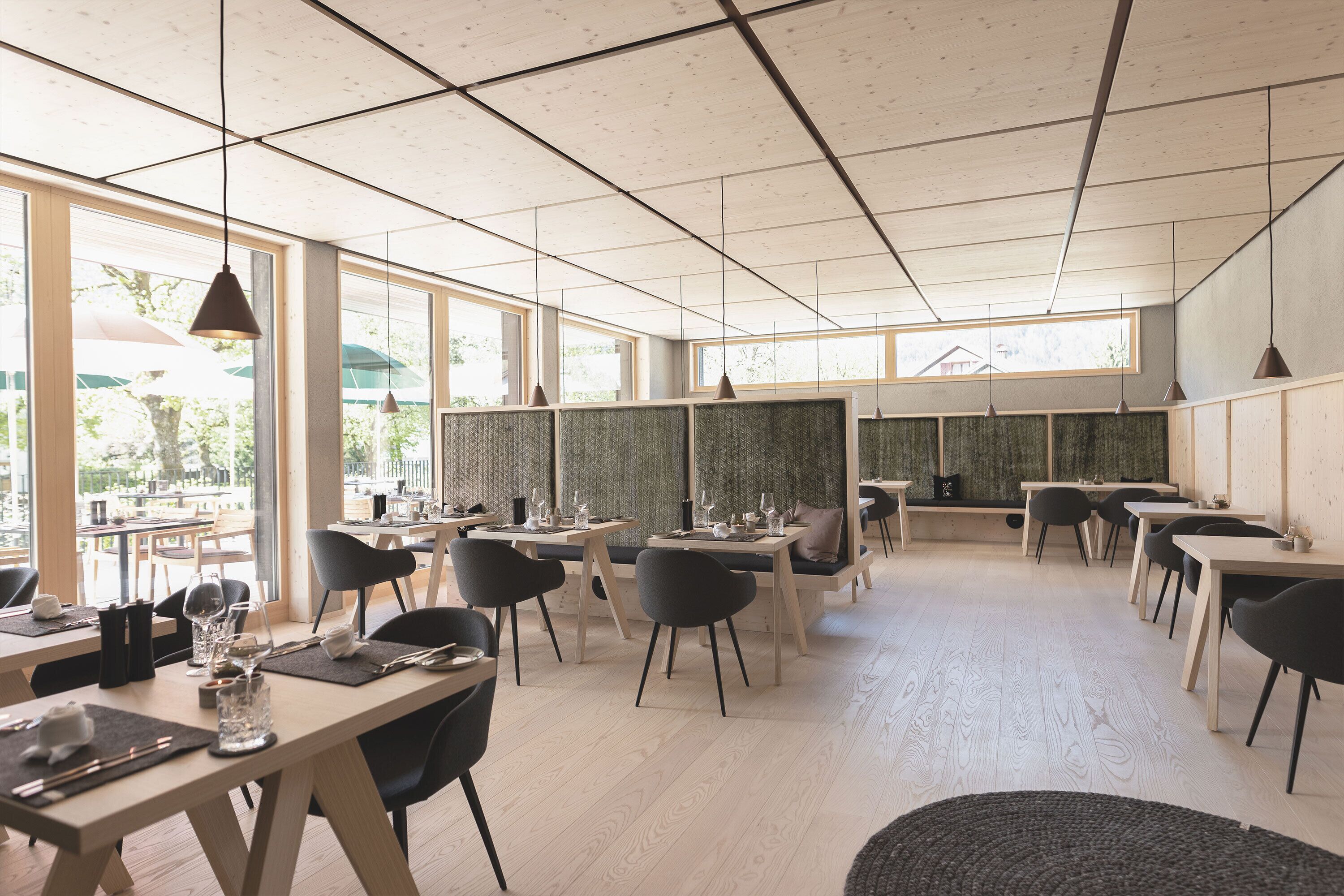 A modern dining area with multiple tables and chairs, large windows, and a high ceiling with exposed beams.