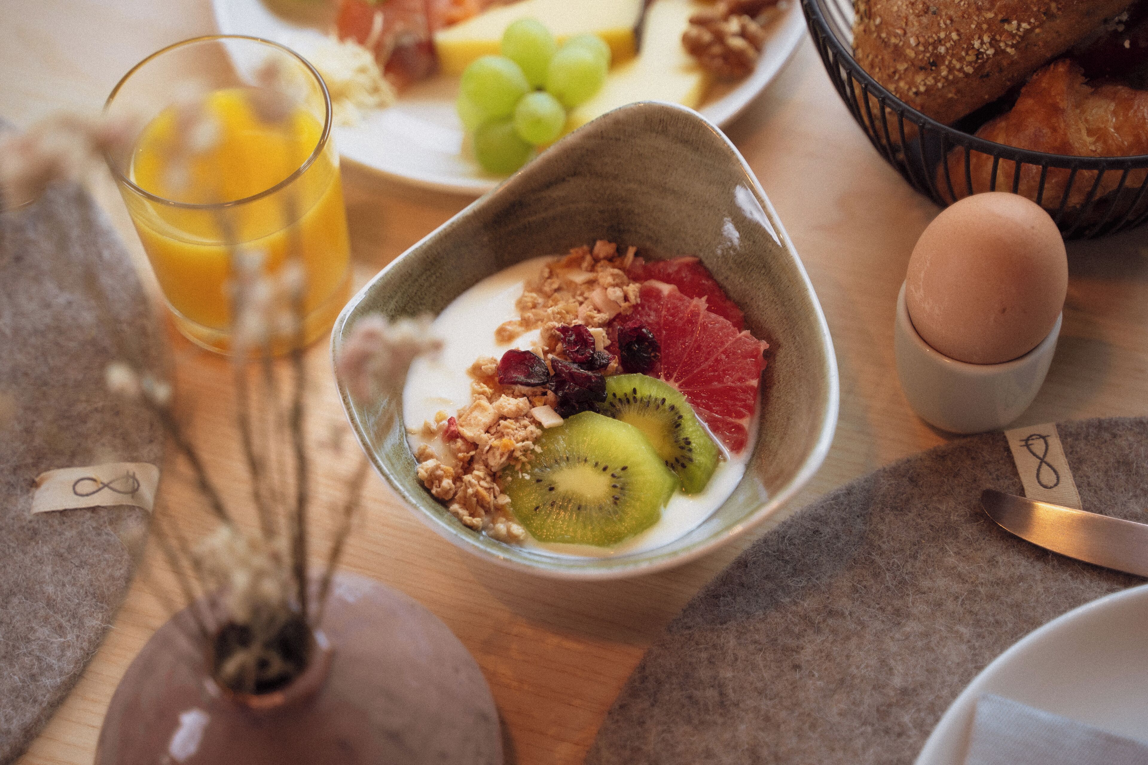 Un desayuno con un tazón de yogur cubierto de granola, kiwi y frutos rojos, un vaso de jugo de naranja y una cesta de pan.