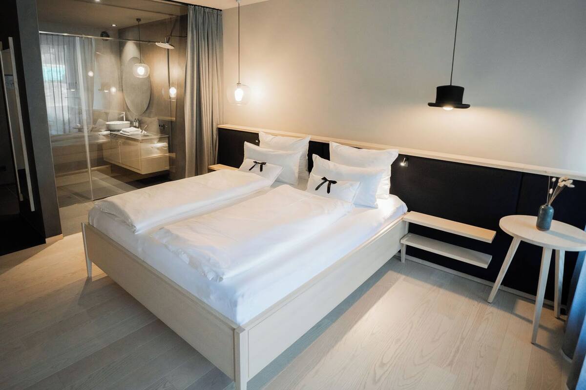 A modern hotel room with a large bed, a bedside table, a mirror, and a small round table with a vase.
