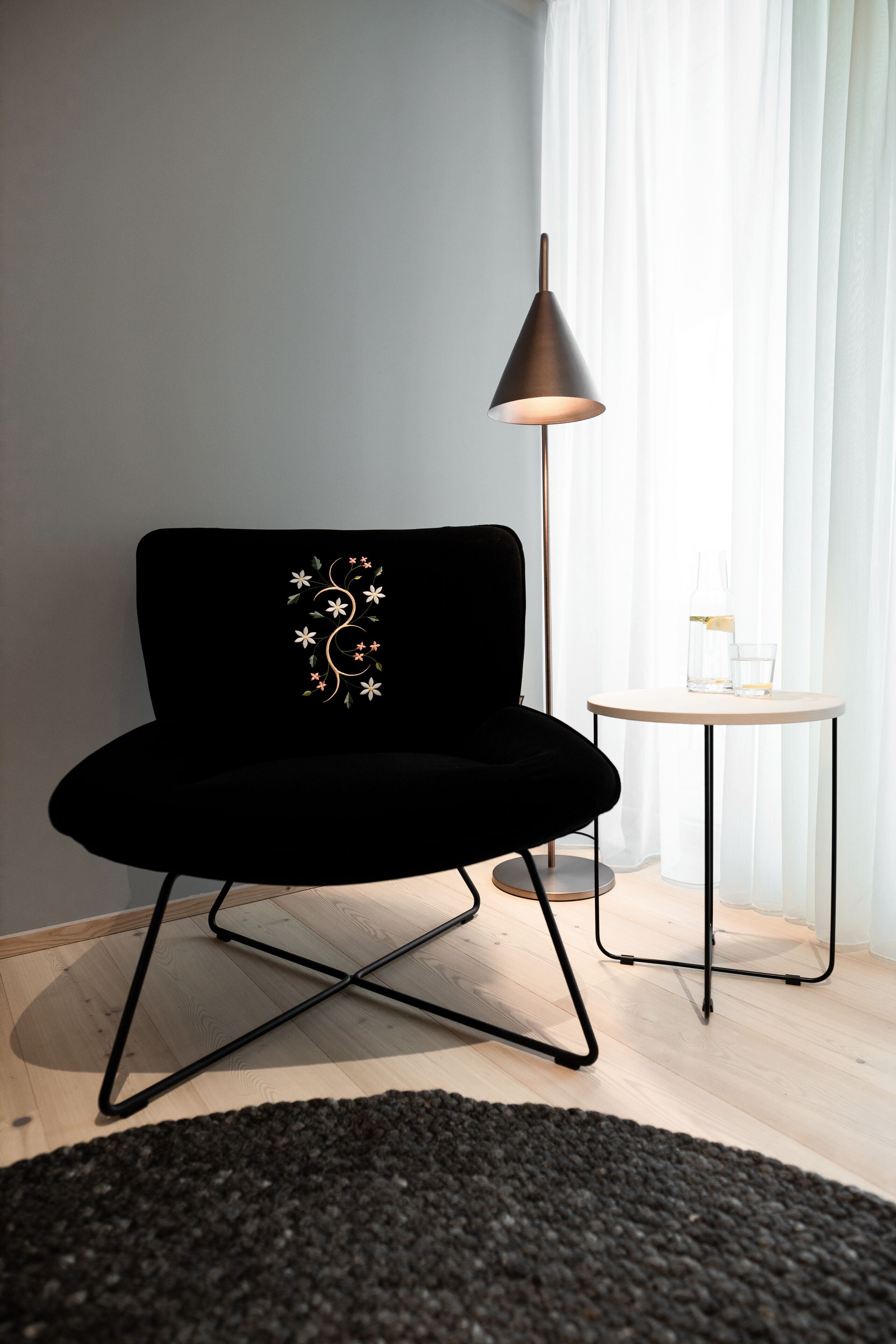A black chair with a floral design, a small round table with a bottle and glass, and a floor lamp.