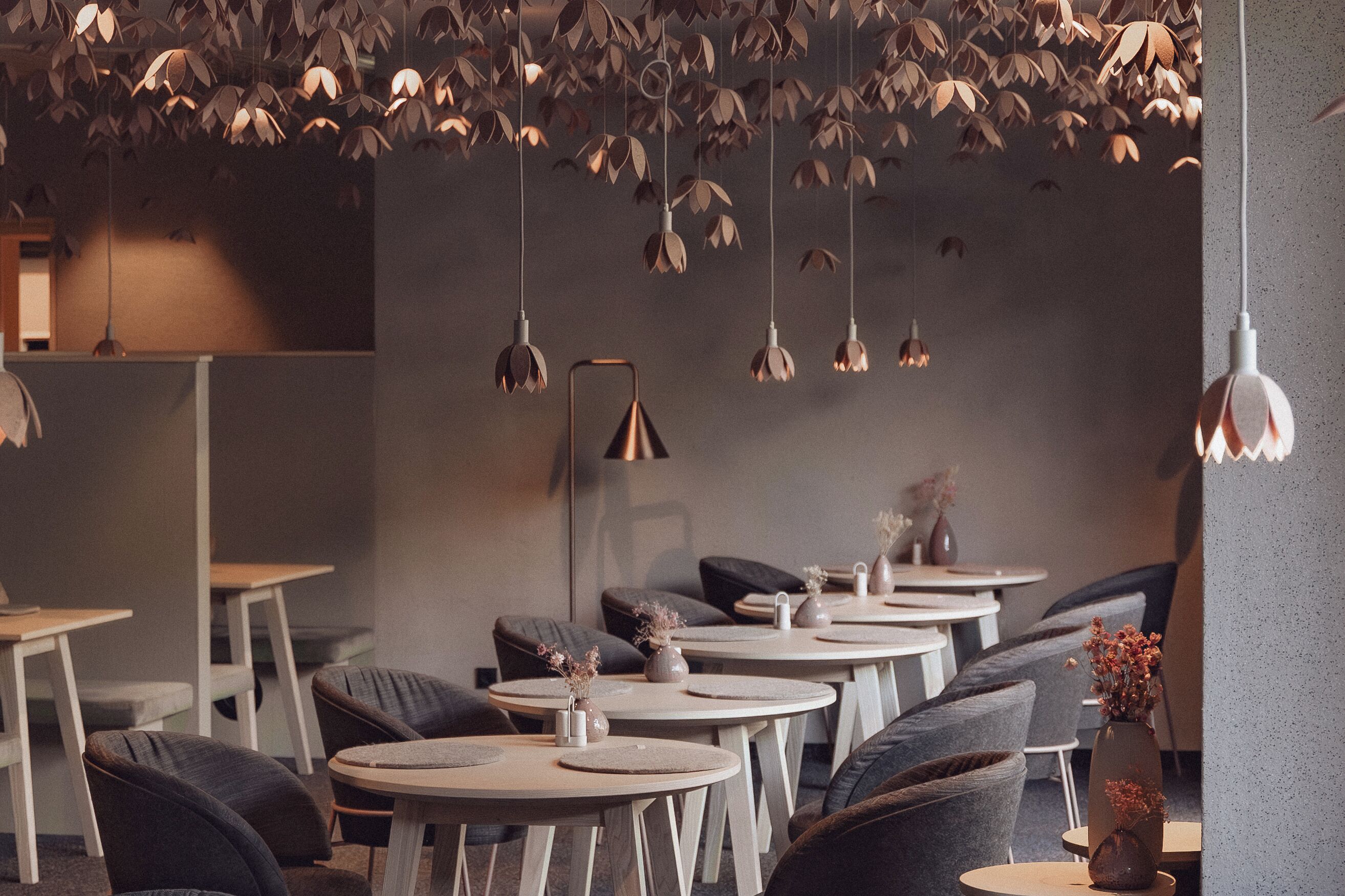 A modern interior with round tables and dark sofas, featuring hanging leaf-shaped lights and small vases with dried flowers.