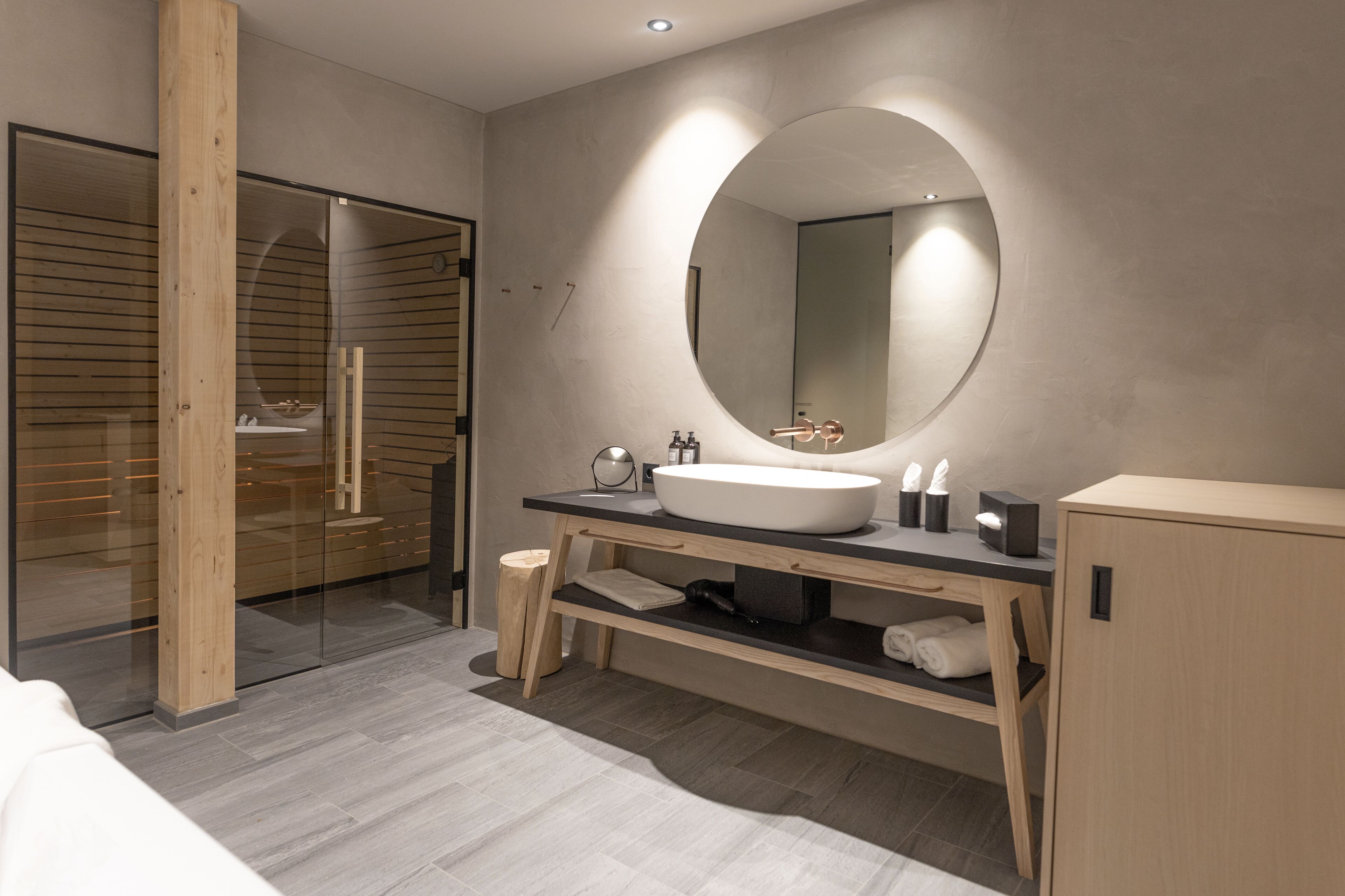A modern bathroom with a large oval mirror, a wooden vanity, and a glass shower enclosure.