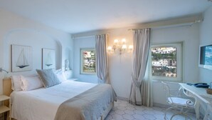 Ein Schlafzimmer mit einem Bett, einem Schreibtisch und einem Stuhl. Zwei Fenster mit Vorhängen, eine Chandelieber und Kunstwerke an den Wänden.