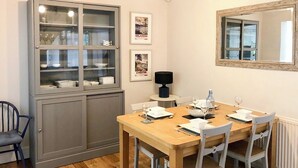 Un comedor con una mesa de madera dispuesta para cuatro personas, rodeada de sillas blancas. A la izquierda, un aparador con puertas de vidrio, y un espejo cuelga en la pared sobre la mesa.