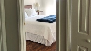 Hotellihuone, jossa on puulattia, valkoisilla lakanoiden ja sinisellä peitolla varustettu sänky, yöpöytä lampun kanssa ja seinällä oleva taulu.