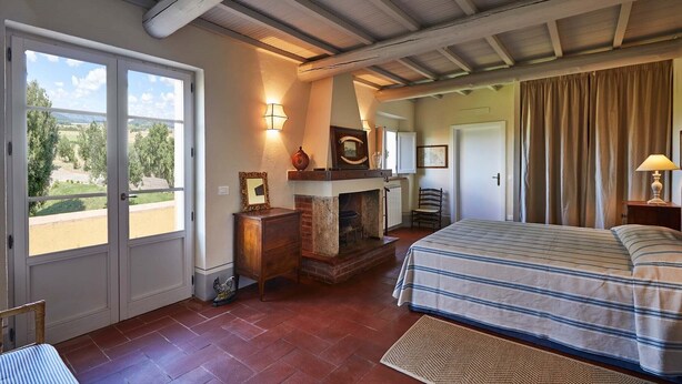 Un dormitorio con chimenea, una cama con ropa de cama a rayas, vistas por una ventana y una puerta que da a otra habitación.