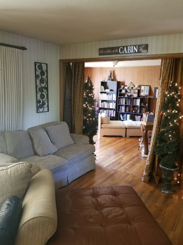 A cozy living room with a sofa, a coffee table, and a bookshelf. There are two Christmas trees and a sign that reads "Life is better at the CABIN."