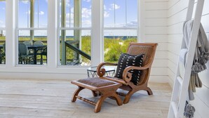 A wooden deck with a wicker chair and ottoman, large windows with a view of the beach, and a white railing.