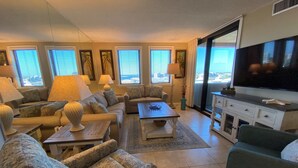 A living room with a sofa, armchairs, a coffee table, and a TV. There are windows with blinds, a lamp, and a cabinet. - Sunset Jetty View! Enjoy one free beach chair set up and umbrella each day! (Destin)