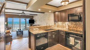 A modern kitchen with granite countertops, stainless steel appliances, and dark cabinetry. It features a large island with a sink, a ceiling fan, and a view of the ocean through large windows. - Direct Beach Front, High End Vacation,Snow Bird Special WINTER 2027 (St. Augustine)