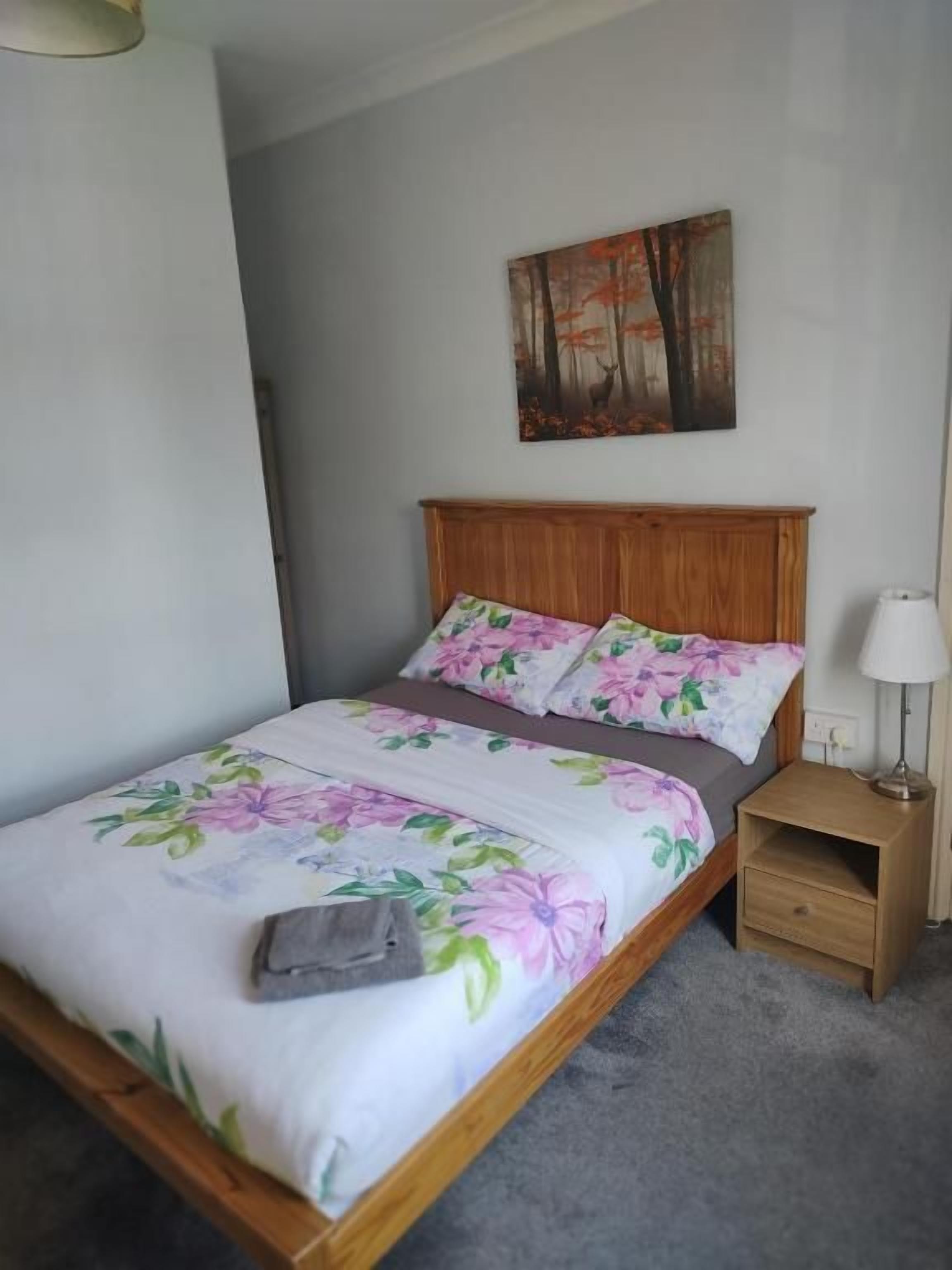 A bedroom with a wooden bed, floral bedding, a nightstand with a lamp, and a painting on the wall.