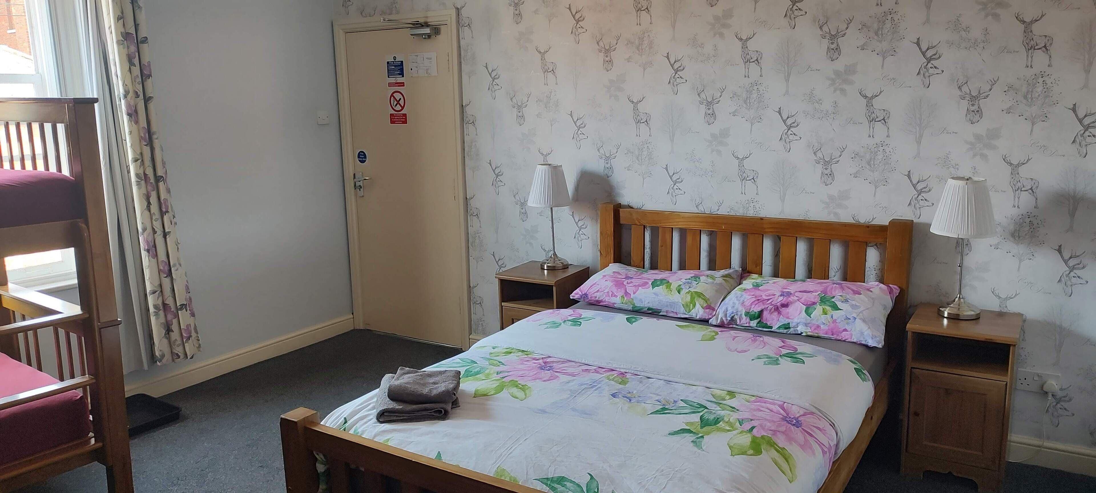 Ein Schlafzimmer mit einem hölzernen Bett, floralem Bettzeug, zwei Nachttischlampen, einer Tür mit Hinweisschildern und einem Fenster mit Vorhängen.