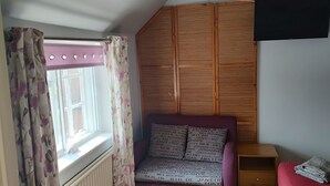 A room with a window, a wooden wardrobe, a purple sofa, and a small table with a folded towel. - Reindeer Accommodation (Southwell)