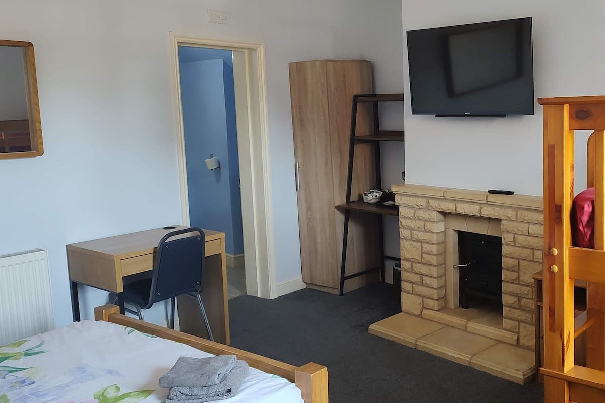 A bedroom with a wooden bed, a desk, a chair, a wardrobe, a television, and a fireplace.