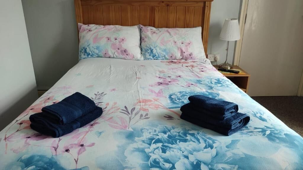 A bed with floral bedding, two rolled towels, a wooden headboard, a bedside lamp, and a nightstand with a lamp.