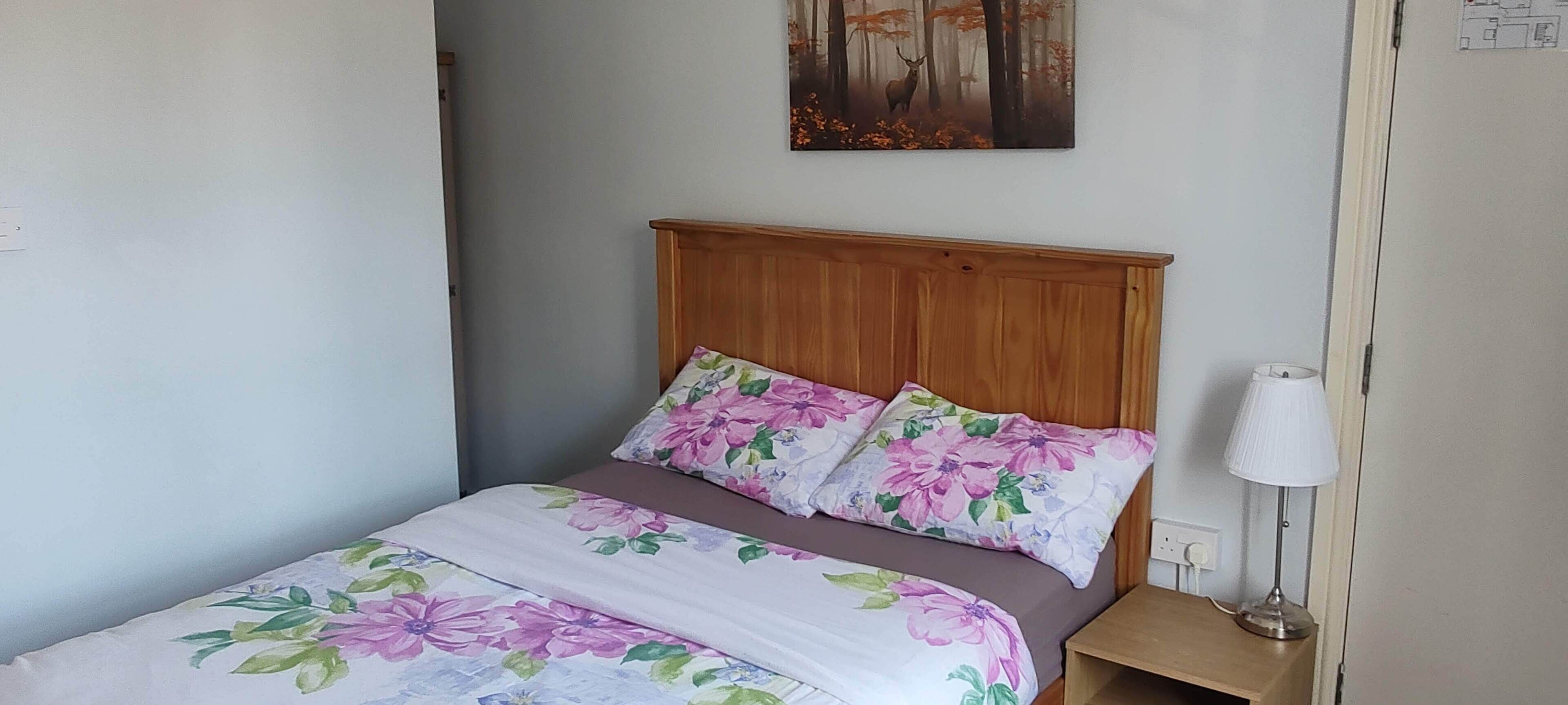 Un dormitorio con cabecera de madera, ropa de cama floral, una lámpara de noche y un cuadro en la pared.