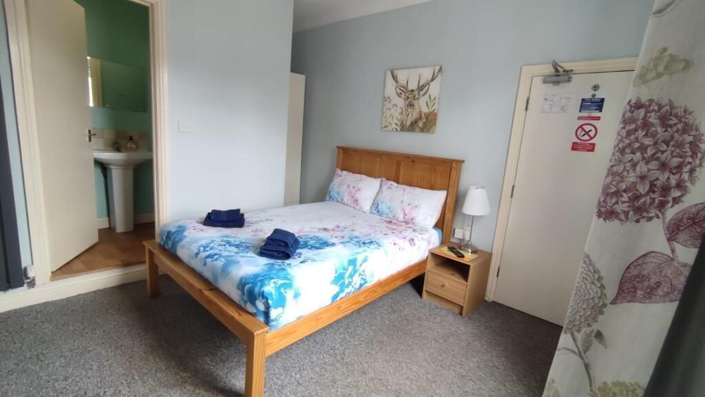 A bedroom with a wooden bed, a nightstand, a wall-mounted lamp, a bathroom visible through an open door, and a floral curtain.
