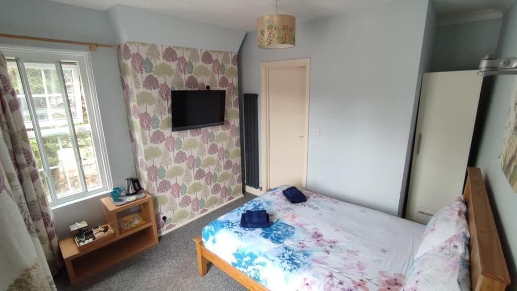 A bedroom with a bed, a television mounted on the wall, a wooden nightstand, and a window with curtains.