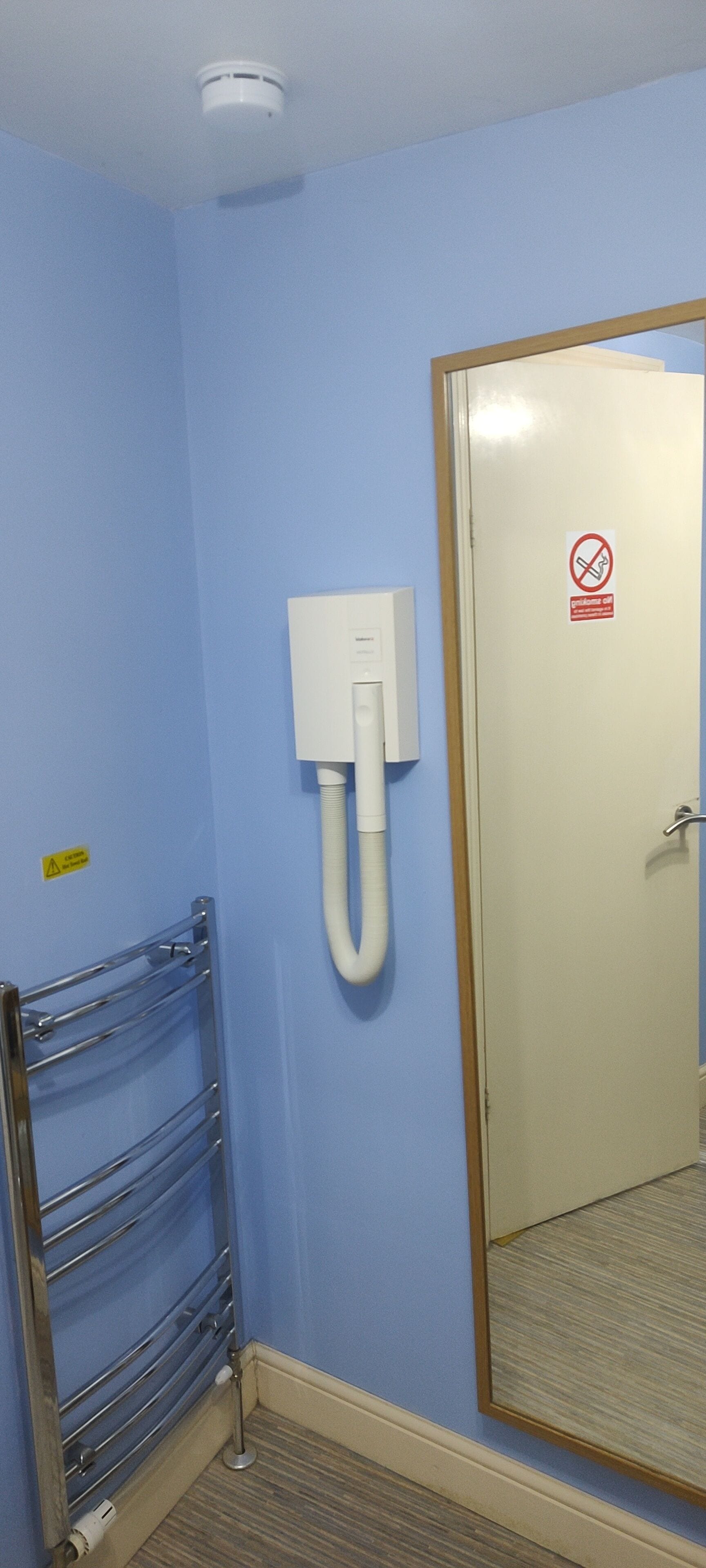 A room with light blue walls, a white door with a no smoking sign, a white electrical box, and a metal towel rack.