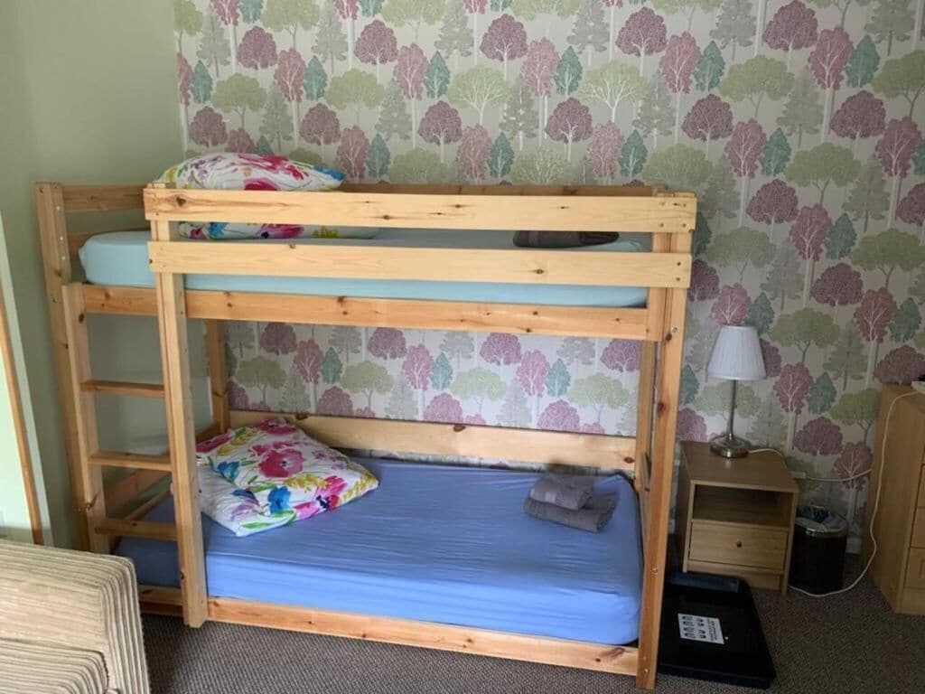 A bunk bed with a blue mattress and floral bedding, a wooden nightstand, a lamp, and a patterned wallpaper background.