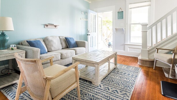 A living room with a light blue wall, a wooden coffee table, a beige sofa with blue cushions, a wicker chair, and a patterned rug.