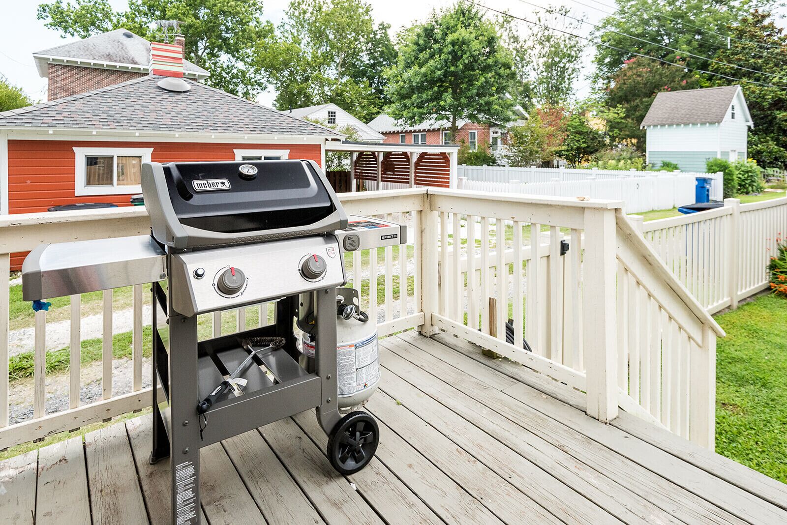 Een draagbare barbecue op een houten terras, met een witte omheining op de achtergrond.