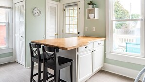A kitchen with a wooden countertop, two black chairs, white cabinets, a clock, and a window with a view of the outside.