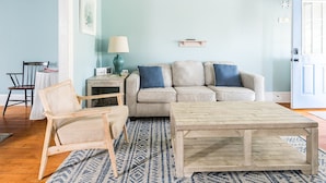 A living room with a light blue wall, a beige sofa with blue cushions, a wooden coffee table, a wicker chair, a small side table with a lamp, and a rug.