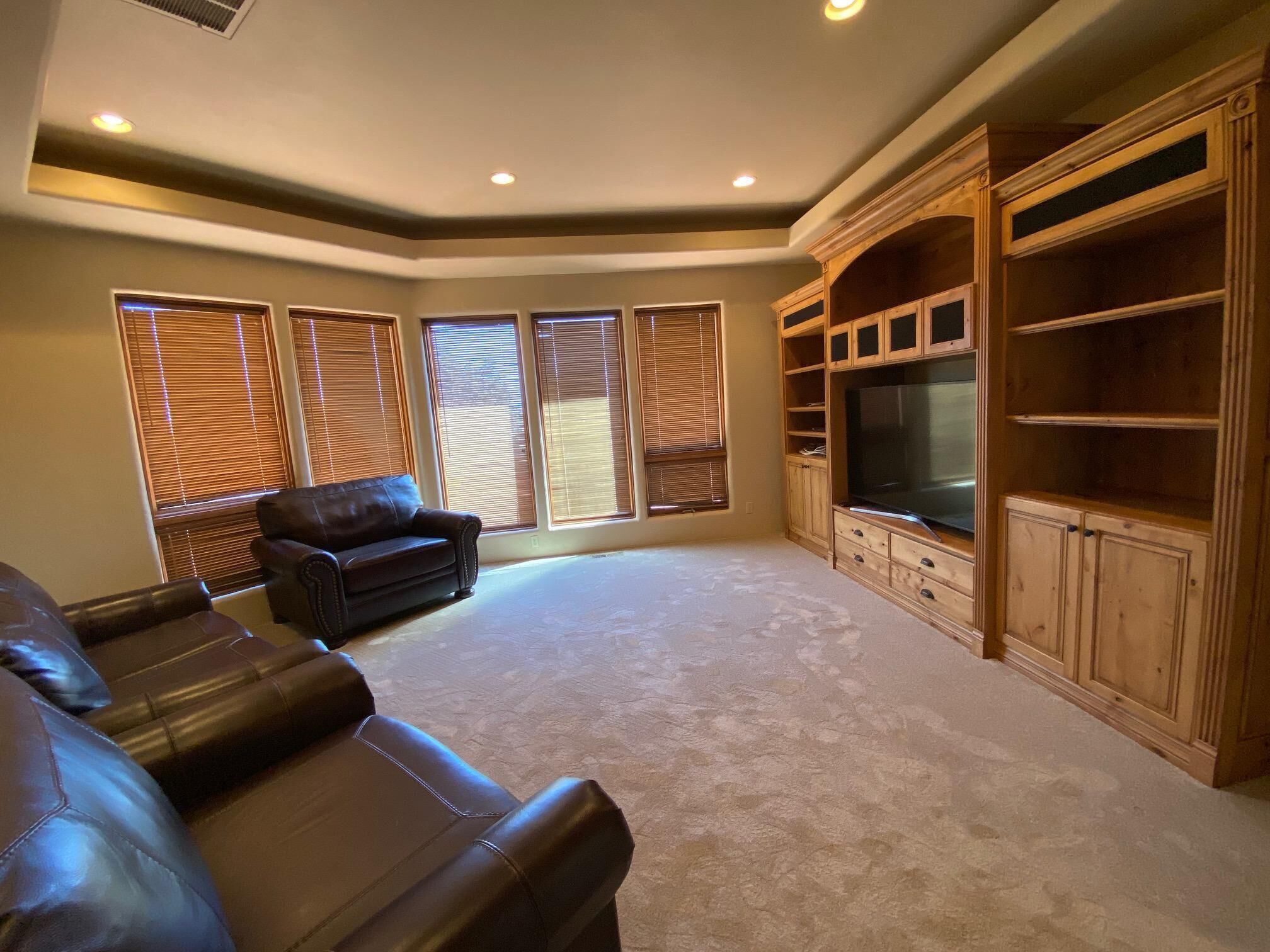 A spacious living room with a leather sofa, built-in entertainment center, and large windows with blinds.