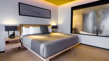 A modern hotel room with a large bed, bedside tables with lamps, a cityscape view through a large window, and a unique wall art piece.