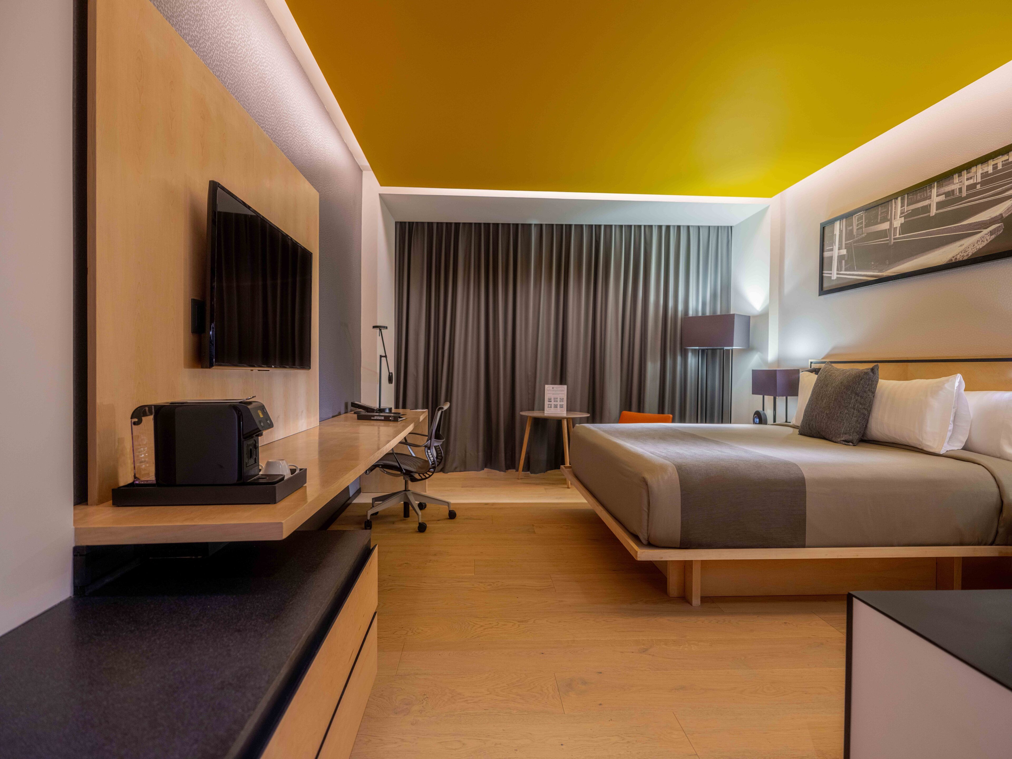 A hotel room with a bed, a desk with a computer, a TV, and a window with curtains.