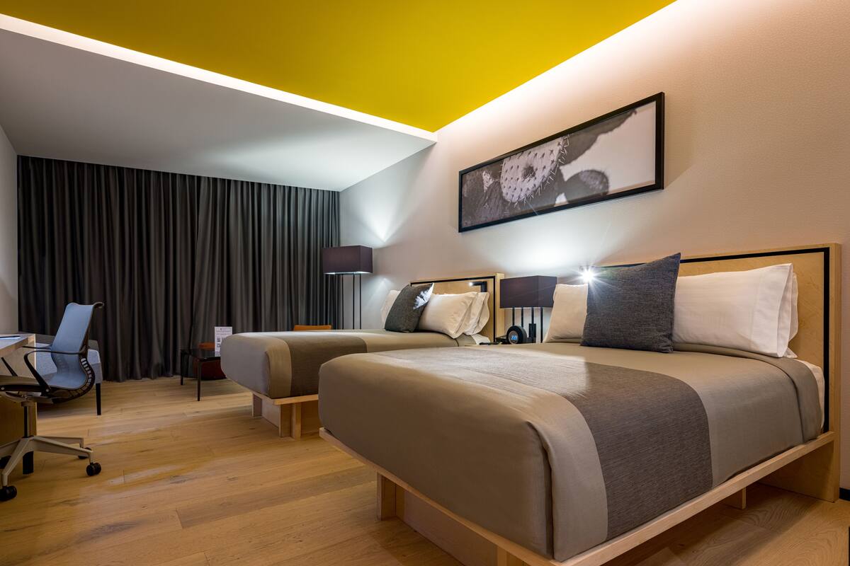 Una habitación de hotel con dos camas, un escritorio, una silla y un ventanal grande con cortinas.