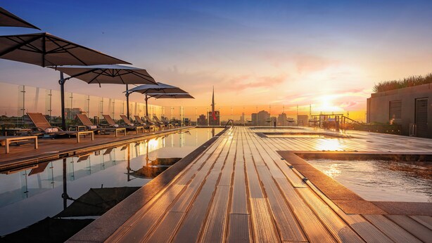 Zona de piscina en la azotea con tumbonas y sombrillas, una terraza de madera y un horizonte urbano de fondo.